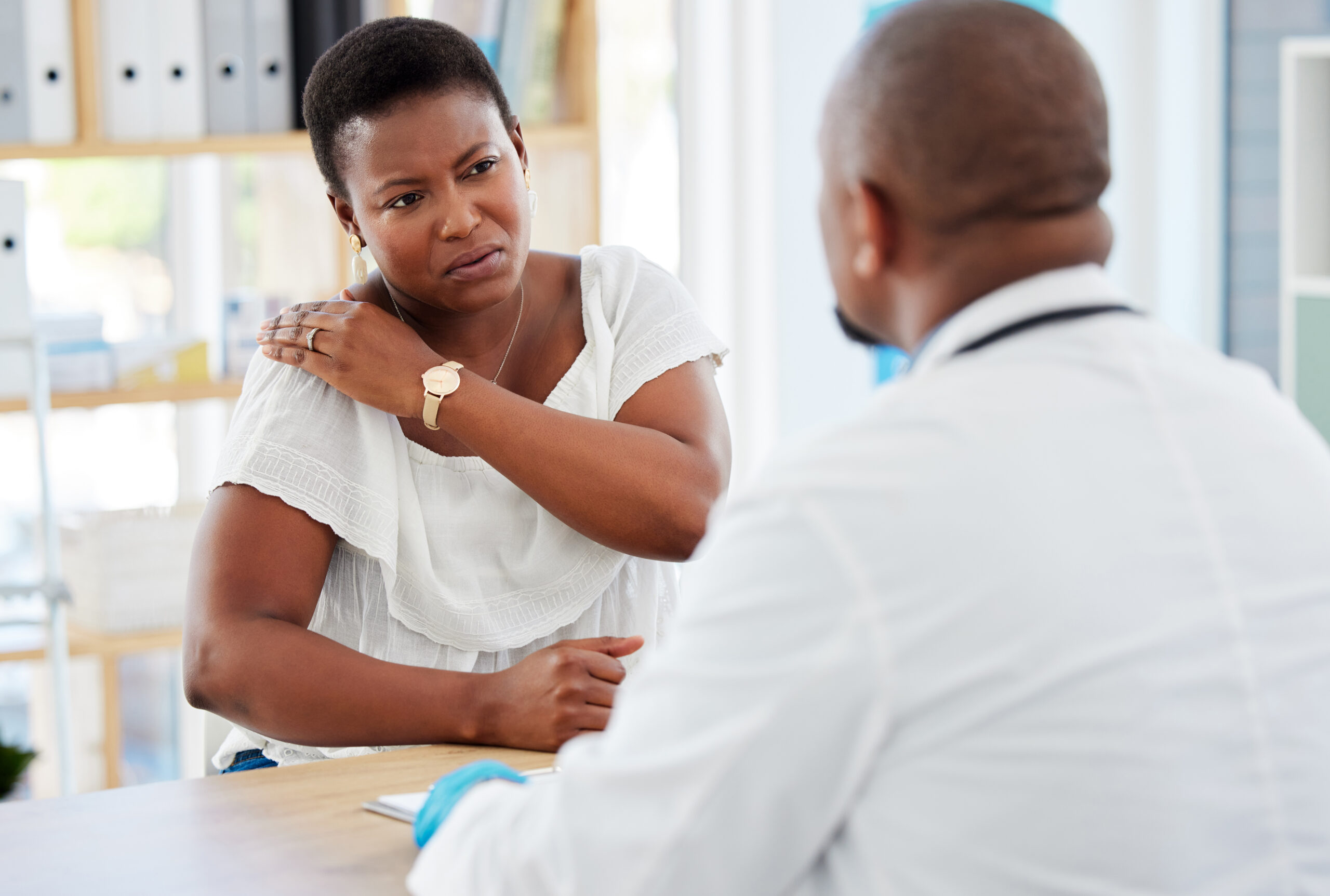 A middle-aged Black woman grips her shoulder and looks like she is in pain as she speaks to a Black physician about shoulder arthroscopy.