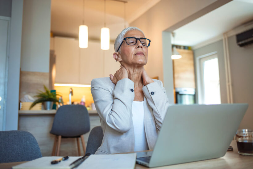 Older woman with gray hair and glasses clutches the back of her neck as she sits at a table near her laptop. She may have spinal stenosis.