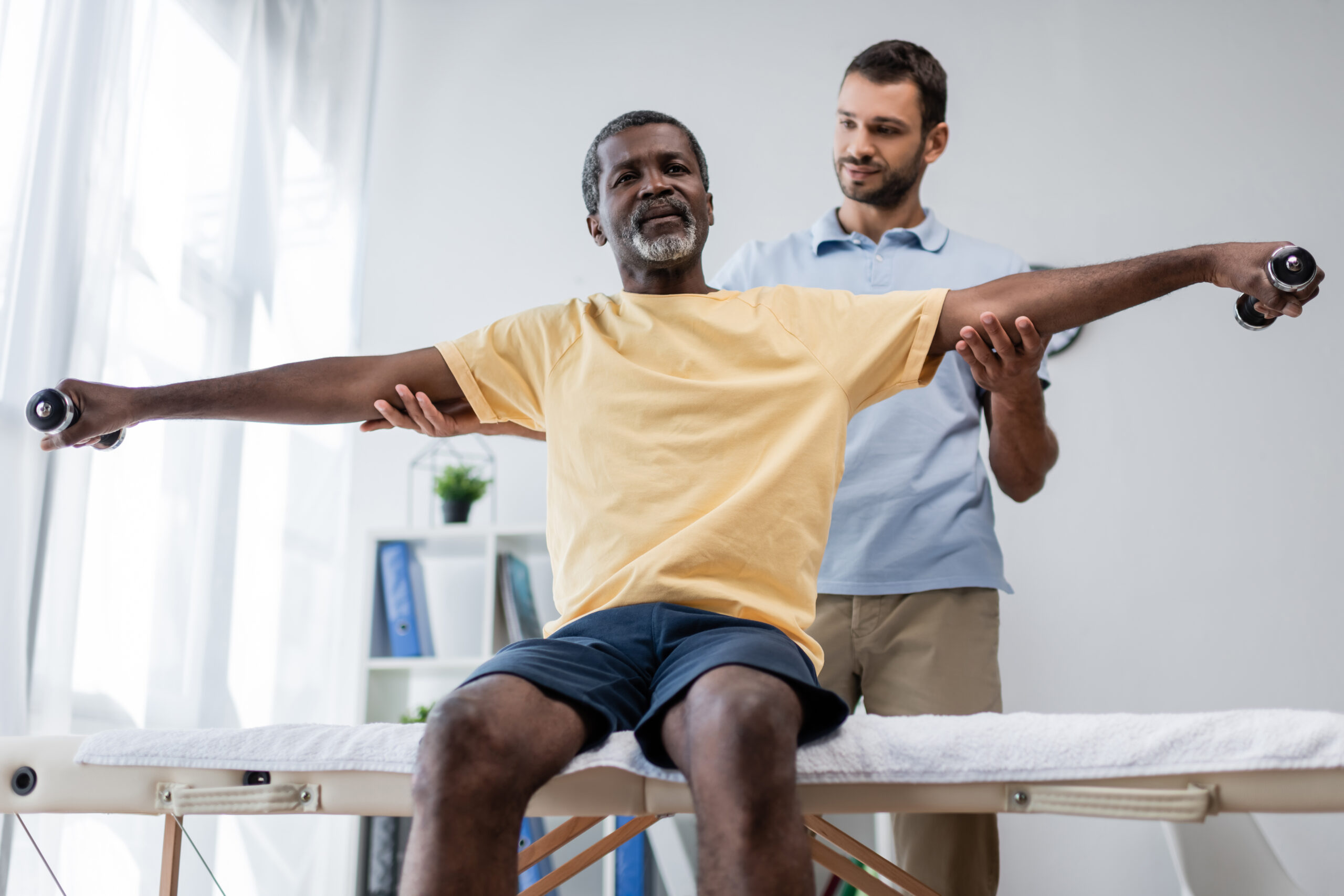 Older black man who received reverse shoulder replacement surgery is holding weights while being examined by a younger male physical therapist